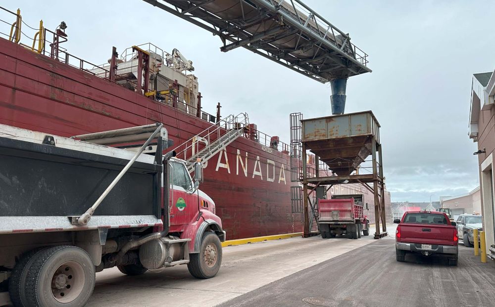 A large red cargo ship with the word “CANADA” painted on its side is docked beside an industrial loading area. Dump trucks and a red pickup truck are lined up on a narrow roadway next to the ship. An overhead conveyor system and a rusty metal chute are positioned to load or unload material between the ship and the trucks. The scene appears busy and industrial, with cloudy skies above.